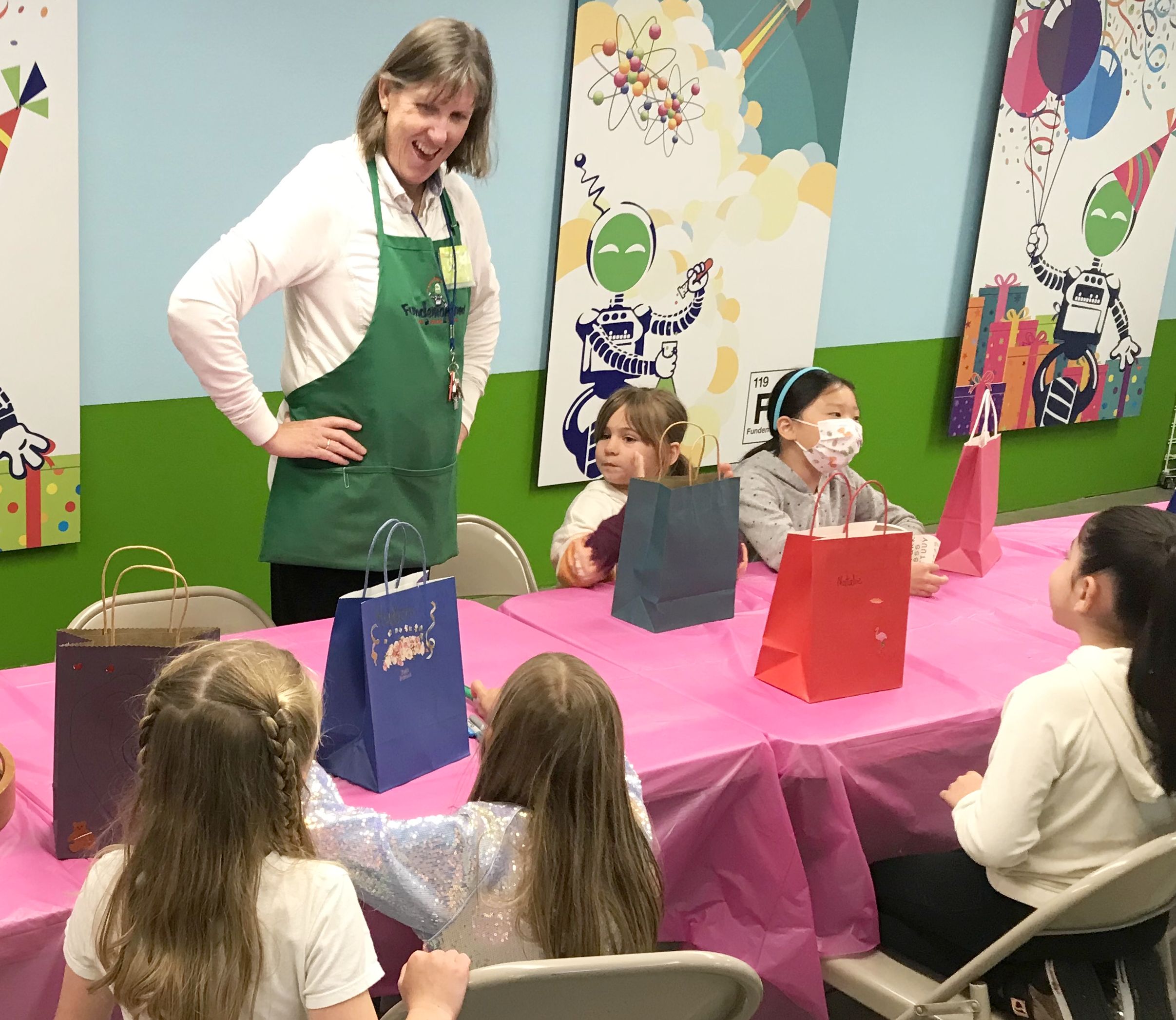 A smiling woman in a green apron oversees a birthday party, standing beside a pink table where children are seated with colorful gift bags, in a room decorated with whimsical party banners."