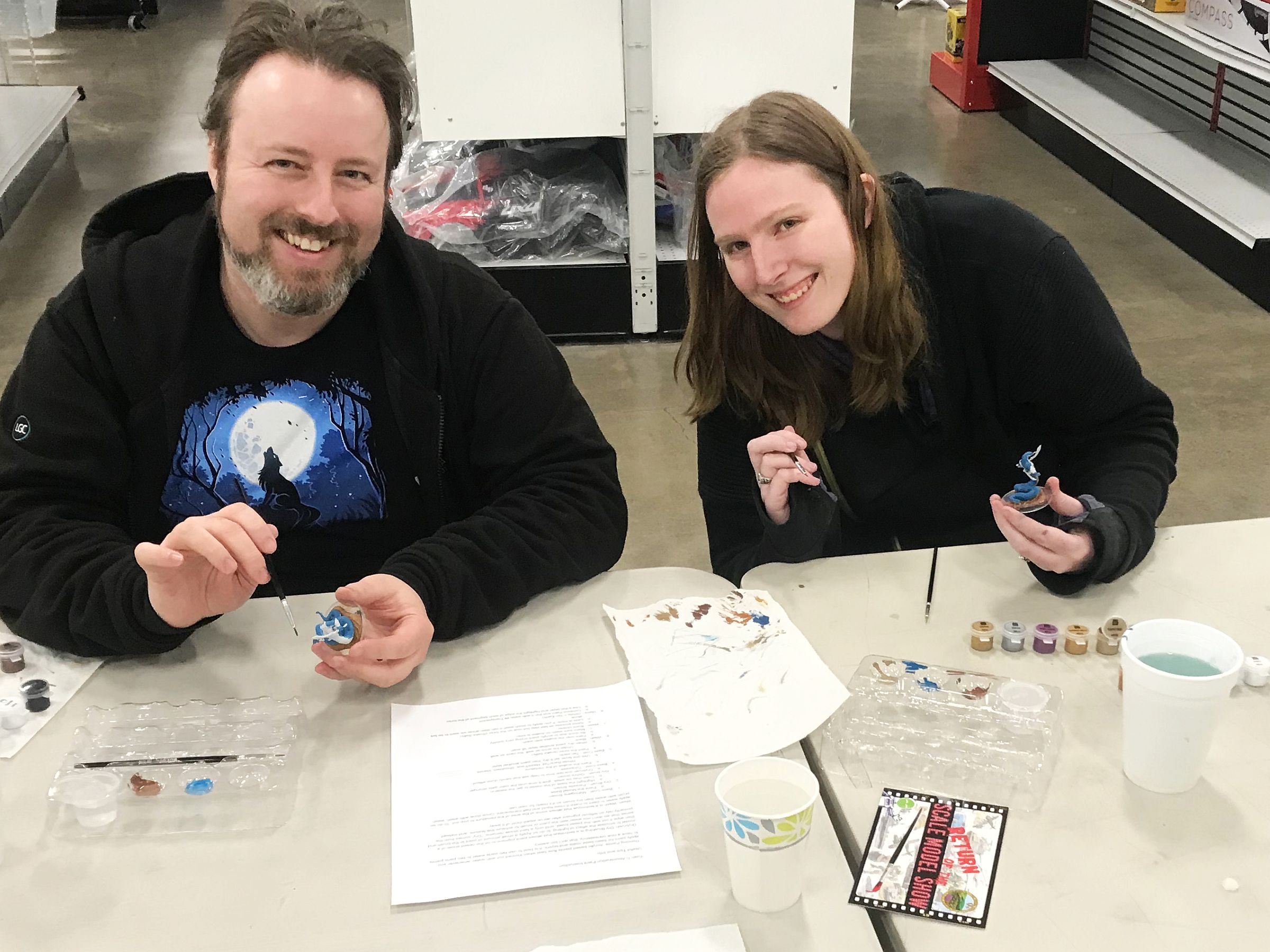 A smiling man and woman sit at a table in a hobby store, happily painting miniature figurines with fine brushes, displaying their artistic skills and shared enjoyment of the craft