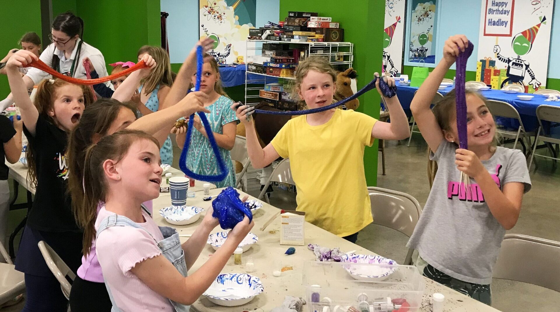 A group of lively children stretch colorful slime between their hands at a science-themed birthday party, with expressions of wonder and joy, as a staff member assists, set against a backdrop of shelves with board games